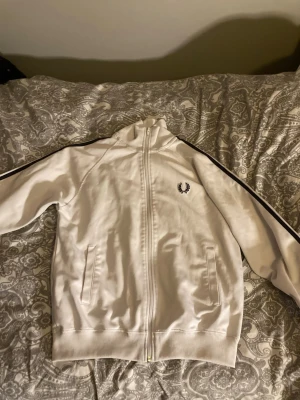 Vit track jacket från Fred Perry - Snygg vit track jacket från Fred Perry med svart logga på bröstet och svarta ränder längs ärmarna. Jackan har dragkedja framtill, ståkrage och två fickor. Tillverkad i ett mjukt och stretchigt material som passar perfekt till streetwear-stilen. Pris går att Diskuteras