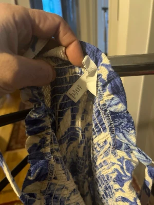 Blåvit blommig klänning i bomull - Supersöt klänning med blått blommönster på vit botten. Gjord i mjuk bomull och har smock upptill för en bekväm passform. Perfekt för varma dagar och har en luftig känsla. Klänningen har en romantisk vibe och är lätt att styla med sneakers eller sandaler.