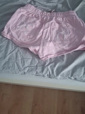 Ljusrosa shorts med snörning och rosetter - Säljer ett par ljusrosa shorts med snörning i midjan och två bakfickor dekorerade med broderade rosetter. Shortsen har en avslappnad passform och är perfekta för varma dagar. Tillverkade i mjuk bomull för extra komfort.
