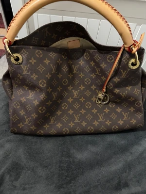 Louis Vuitton Monogram handväska - Säljer en klassisk Louis Vuitton handväska i brunt monogramcanvas med ikoniska LV-loggor. Väskan har en rejäl beige läderrem med röda detaljer och guldfärgade metalldetaljer. Perfekt för dig som vill ha en lyxig och tidlös accessoar.
