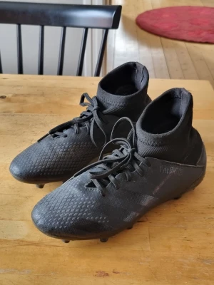 Svarta Adidas Predator fotbollsskor. - Säljer ett par helsvarta Adidas fotbollsskor med hög ankel och snörning. Skorna har en texturerad yta för bättre bollkontroll och är tillverkade i syntetmaterial. Perfekta för dig som vill ha stilrena och funktionella skor på planen.
