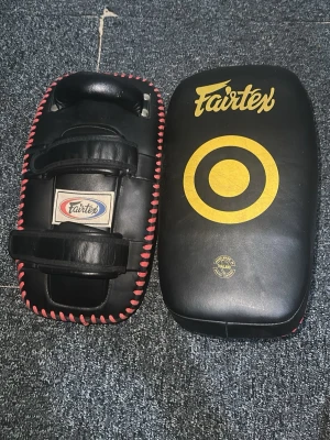 Fairtex svart thaipad med gult tryck - Säljer ett par svarta thaipads från Fairtex med gult tryck och röd snörning runt kanterna. Tillverkade i slitstarkt läder med två justerbara kardborreband och handtag.   Nypris: 2499kr 