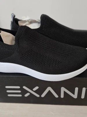Svarta slip-on sneakers från Exani - Svarta slip-on sneakers från Exani med stickad ovandel i mesh för extra andningsförmåga. Skorna har en vit, lättviktig sula och elastisk mudd runt ankeln för enkel påtagning. Perfekta för en sportig och avslappnad stil.