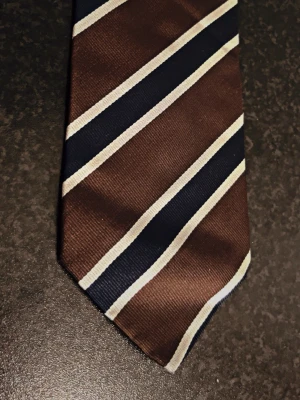 Randig slips från GANT - Snygg slips i brunt med diagonala ränder i marinblått och vitt från GANT. Tillverkad i ett glansigt tyg som ger en stilren look. Perfekt accessoar för att lyfta din outfit med klassiska färger och mönster.