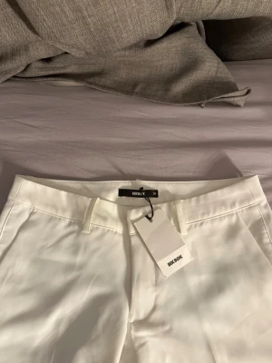 Vita kostymbyxor från Bik Bok - Säljer ett par helt nya  stilrena vita kostymbyxor från Bik Bok i storlek 38. Prislappen finns kvar på byxorna och de  är bootcut , pressveck och hällor för skärp. Materialet känns mjukt och har en lätt glansig finish. Perfekta för en clean och trendig look.
