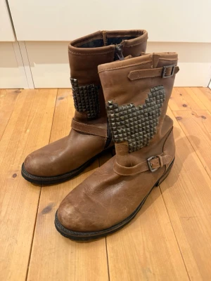 Bruna boots med nitar och hjärtdetalj - Säljer ett par bruna boots i skinnimitation med coola nitar formade som ett hjärta på skaftet. Bootsens design har spännen vid ankeln och upptill samt dragkedja på insidan. Perfekta för dig som gillar en edgy stil. Jag har storlek 39 vanligtvis