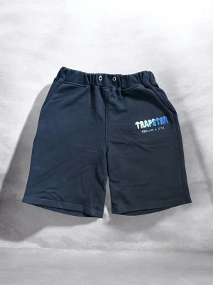 Svarta shorts från Trapstar - Svarta shorts från Trapstar med elastisk midja och två fickor. Tryck i blått och vitt på vänster ben där det står 'Trapstar' och 'Reload & Retry'. Perfekta för chill dagar och streetwear-stil.