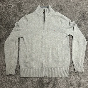 Grå kofta från Tommy Hilfiger - Säljer en stilren grå kofta från Tommy Hilfiger med dragkedja framtill och diskret logga på bröstet. Tröjan har ribbade muddar och hög krage, perfekt för lager-på-lager. Passar till det mesta och är riktigt skön.