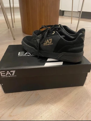 Svarta EA7 Emporio Armani sneakers - Snygga svarta sneakers från EA7 Emporio Armani med gulddetaljer och logga på sidan. Skorna har en mix av mocka och meshmaterial, samt svarta skosnören och robust sula. En liten fläck på 
