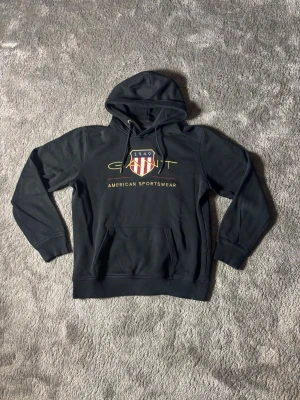 Svart hoodie från Gant - Snygg svart hoodie från Gant med broderad logga och texten 'American Sportswear' på bröstet. Klassisk huva med dragsko och stor magficka framtill. Tillverkad i mjukt material, perfekt för en avslappnad stil.