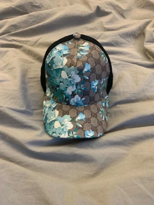 Gucci keps - Snygg keps från Gucci med klassiskt GG-mönster och blå blommor över hela kepsen. Skärmen är böjd och baksidan har svart mesh för extra ventilation. Justerbar rem bak för perfekt passform. Materialet är en mix av canvas och mesh.