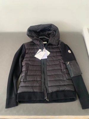 Svart Cardigan från Moncler - Snygg svart helt ny Cardigan från Moncler med quiltad front och huva. Ärmarna är stickade i mjukt material och har en Moncler-logga på vänster ärm samt en liten ficka med dragkedja. Jackan har två framfickor med knappar och dragkedja framtill.