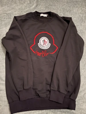 Svart stickad tröja från Moncler - Svart stickad tröja från Moncler med stort broderat logotypmärke i vitt och rött på bröstet. Tröjan har rund halsringning, ribbade muddar och är tillverkad i 100% bomull för en skön och avslappnad känsla.