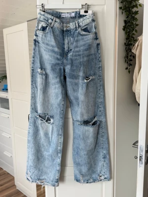 Ljusa baggy jeans med slitningar - Säljer ett par ljusblå baggy jeans med stora slitningar på knäna och fransiga benslut. Jeansen har hög midja, fem fickor och raka ben. Perfekta för en avslappnad och trendig look.