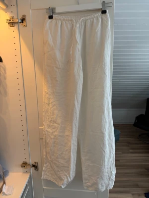 Vita vida linnebyxor med dragsko - Säljer ett par vita linnebyxor med avslappnad passform och dragsko i midjan. Byxorna har raka ben och elastisk midja för extra komfort. Perfekta för en chill och stilren look under varma dagar. Dom är lågmidjade och passar nån som är 170