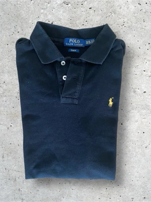 Svart långärmad piké från Polo Ralph Lauren - Klassisk svart långärmad pikétröja från Polo Ralph Lauren med gul broderad logga på bröstet. Skjortan har krage, knappar i halsen och är gjord i mjuk bomull. Perfekt för en clean och stilren look. Storleken är XL men tror de är xl i barnstorlek. Skulle säg att den passar dem som är 170-175