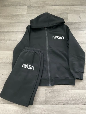Svarta NASA mjukisbyxor - Säljer ett par svarta mjukisbyxor från NASA med vit logga på vänster ben. Byxorna har resår i midjan och är gjorda i mjukt, bekvämt material. Perfekta för chill dagar eller när du vill ha en avslappnad look.