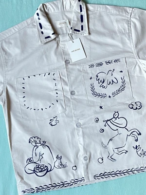 Vit skjorta med blå illustrationer - Unik vit skjorta från Ame Leon Dore med handritade blå illustrationer av figurer, djur och natur. Skjortan har två bröstfickor, klassisk krage med blå detaljer och knäppning framtill. Tillverkad i mjuk bomull och har en loose passform.