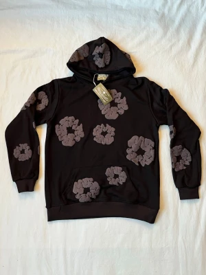 Svart hoodie med gråa blommor - Svart hoodie från Denim Tears med stora gråa blommönster över hela plagget. Klassisk huva och magficka framtill. Tillverkad i mjukt material som känns skönt mot huden. Perfekt för dig som vill sticka ut med en unik och trendig stil.