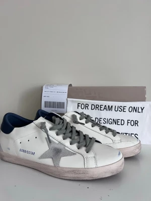 Golden Goose vita sneakers med stjärna - Snygga vita sneakers från Golden Goose med grå mockastjärna på sidan, grå skosnören och mörkblå häl. Skorna har GGDB/SSTAR-tryck och en vintage-inspirerad sula. Perfekta för dig som gillar en avslappnad men trendig stil.