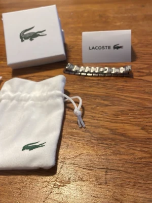 Silverfärgat armband från Lacoste - Snyggt armband från Lacoste i silverfärgat rostfritt stål med länkdesign och diskreta mönstrade detaljer på varje länk. Kommer med original ask och tygpåse med Lacoste-logga. Perfekt accessoar för dig som gillar stilrena smycken med sportig touch.