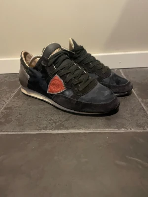 Philippe Model svarta sneakers - Snygga svarta sneakers från Philippe Model med mörkblå kamouflagemönster och en röd detalj på sidan. Skorna har svarta skosnören, mockadetaljer och vit sula. Perfekta för dig som vill ha en unik och trendig look.