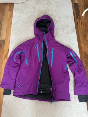 Lila vindjacka från Norrøna med huva - Snygg lila vindjacka från Norrøna med turkosa dragkedjor och flera praktiska fickor. Jackan har justerbar huva, kardborre vid ärmslut och tumhål. Materialet är slitstarkt och vattenavvisande, perfekt för blåsiga dagar. Logga på ärm och rygg.