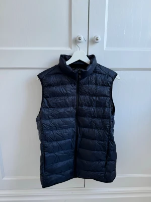 Svart dunväst med hög krage - Snygg blå dunväst med quiltad design och hög krage från uniqlo. Västen har dragkedja framtill och är lätt vadderad för extra värme. Perfekt att slänga över en hoodie eller tröja för en chill look.