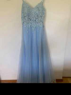 Ljusblå långklänning med spetsdetaljer - Säljer en ljusblå långklänning med smala axelband och v-ringning. Överdelen har vacker spets och broderade detaljer, medan kjolen är i flera lager tyll för en drömmig look. Perfekt för bal eller festliga tillfällen. Nypris 1800