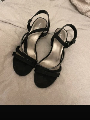 Svarta klackar med klack Glitter - Stilrena svarta sandaletter från Glitter med bred klack och smala remmar. En av remmarna är dekorerad med små glittrande stenar för extra detalj. Sandalerna har öppen tå och justerbart spänne vid ankeln. Materialet är mockaimitation.