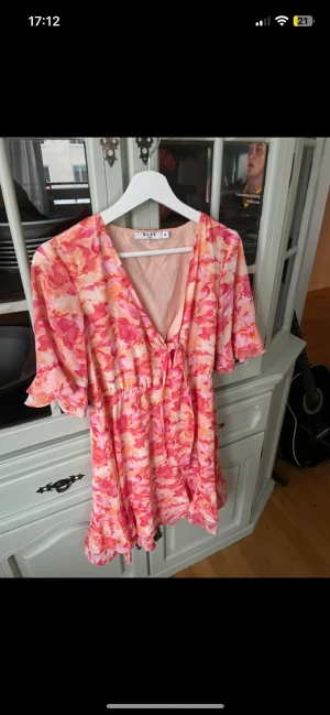 Blommig omlottklänning från NA-KD - Supersöt kort omlottklänning från NA-KD i blommigt mönster med rosa, orange och vita toner. Klänningen har volangdetaljer, v-ringning och korta ärmar. Perfekt för sommarens alla tillfällen och ger en fräsch och färgglad look.