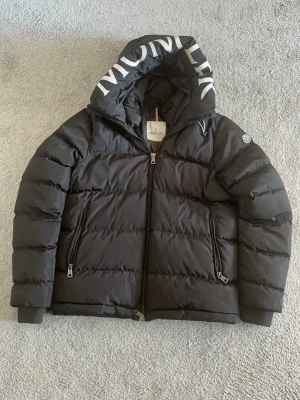 Svart dunjacka från Moncler med huv -  Snygg svart dunjacka från Moncler med stor huva där Moncler-loggan är tryckt i vitt. Jackan har två dragkedjeförsedda fickor, quiltad design och klassisk Moncler-patch på ärmen. Perfekt för kalla vinterdagar och riktigt trendig stil.