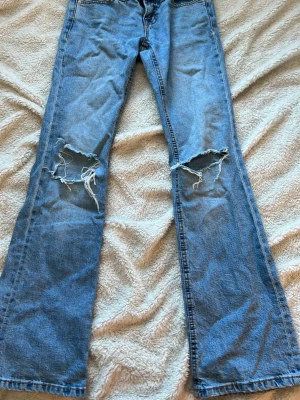 Blå slitna bootcut jeans - Säljer ett par ljusblå bootcut jeans med stora slitningar över båda knäna. Jeansen har klassisk femficksdesign och är tillverkade i denim. Perfekta för en avslappnad och trendig look. 