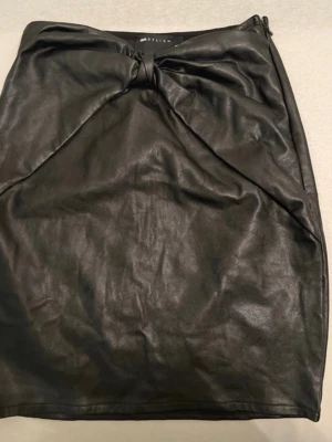 Svart skinnkjol i kort modell - Snygg svart kort kjol i skinnimitation med enkel och stilren design. Kjolen har en rak passform och är perfekt för dig som gillar en edgy look. Passar till både t-shirt och stickad tröja.