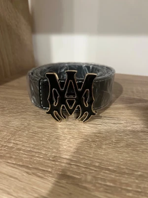 Svart bälte med stor metalllogga - Säljer ett svart bälte med bred rem och en stor, iögonfallande metallspänne med unik logotyp. Bältet har vita sömmar längs kanterna och är gjort i skinnimitation med slät finish. Perfekt för att lyfta vilken outfit som helst.