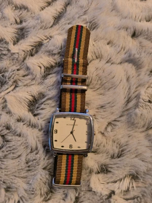 Burberry klocka med randigt band - Snygg Burberry klocka med fyrkantig silverfärgad boett och vit urtavla. Klockan har ett randigt textilband i beige, rött, grönt och svart med metallspänne. Perfekt accessoar för dig som gillar klassisk men ändå unik stil.