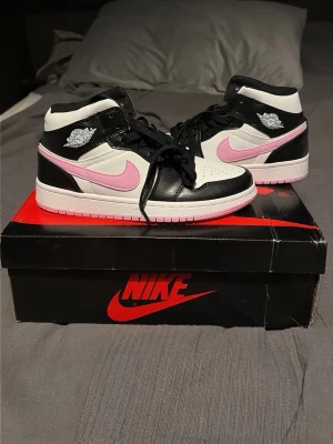Nike Air Jordan 1 Mid – Rosa Swoosh - Nike Air Jordan 1 Mid sneakers med svart, vit och rosa färgkombination. Klassisk design med perforerad tå, rosa swoosh och Wings-logga på sidan. Tillverkade i läder med svarta skosnören och vit/rosa yttersula. Perfekt för dig som vill sticka ut.