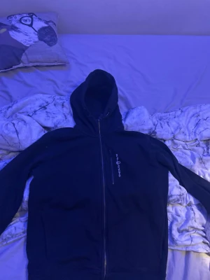 Svart hoodie från Sail Racing - Säljer en svart hoodie från Sail Racing med dragkedja framtill och en bröstficka med dragkedja. Hoodien har justerbar huva med snören och logga tryckt på bröstet. Perfekt för dig som gillar sportig och clean stil.