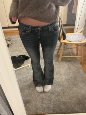 Lågmidjade jeans - Säljer ett par mörkblå bootcut jeans med snygga slitningar och vita kontrastsömmar. Bakfickorna har stora broderade detaljer i vitt som ger en unik look. Jeansen har låg midja och klassisk femficksmodell. Storlek M på ginas 14+ avdelning så passar S