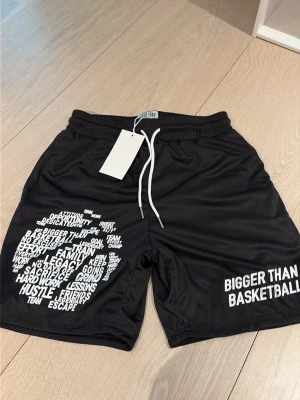 Svarta basketshorts Bigger Than basketball  - Säljer ett par svarta basketshorts från Bigger Than med vita tryck och ord på framsidan. Shortsen har elastisk midja med vit snörning och är gjorda i ett lätt, ventilerande material som passar perfekt för sport och träning. Den är S i killstorlek och M i tjejstorlek. 