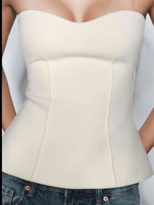 Krämvit axelbandslös korsettopp - Snygg krämvit korsettopp med axelbandslös design och markerad midja. Toppen har en stilren och figurnära passform som ger en modern siluett. Perfekt att styla med jeans eller kjol för en trendig look. Helt oanvänd och prislapp kvar! Lite för stor för mig.