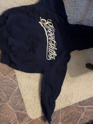 Mörkblå Silverticket hoodie - Mörkblå hoodie från Silverticket med stor vit logga i snyggt skriven text på bröstet. Klassisk känguruficka framtill och huva. Tillverkad i mjukt material, perfekt för chill dagar. Långärmad och ribbade muddar vid ärmslut och nederkant.