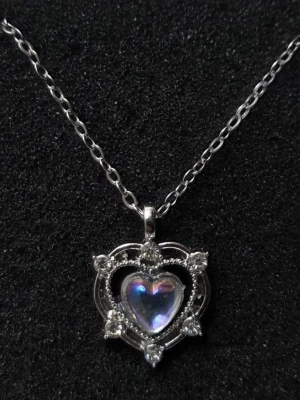 Halsband med hjärthänge i silverfärg - Snyggt halsband med tunn silverfärgad kedja och ett hänge format som ett hjärta. Hänget har en glittrande, pärlemorskimrande sten i mitten och är dekorerat med små gnistrande stenar runt om. Perfekt accessoar för att lyfta din stil.  Detaljer: Design: Hjärtformad krona med skiftande sten Huvudmaterial: Legering i silverton               Sten: Hjärtformad opaliserande sten Färg: Silver med skimrande vit/rosa/lila sten          Kedjelängd: 45 cm Skick: Helt nytt / Oanvänt