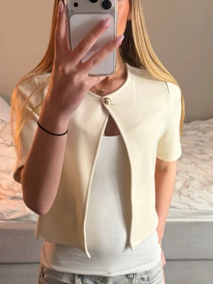 Kortärmad cremevit kofta - Snygg cremevit kofta med korta ärmar och en dekorativ knapp framtill. Koftan har en öppen design och är tillverkad i ett mjukt, strukturerat material som ger en clean och modern look. Perfekt att slänga över ett linne för en fräsch outfit.