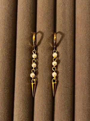 Guldörhängen med pärlor och spikes - Snygga örhängen i guldton med fyra små vita pärlor på rad och en spetsig spike-detalj längst ner. Hängande modell med klicklås. Perfekt för dig som gillar en mix av elegant och edgy stil.