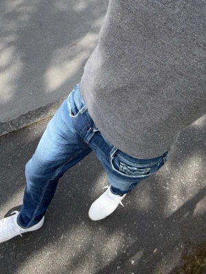 Blåa Replay jeans med slitningar - Säljer ett par blåa jeans från Replay med snygga slitningar och tvättade detaljer. Klassisk femficksmodell med raka ben och låg midja. Jeansen har vita kontrastsömmar och är gjorda i mjukt denimtyg som sitter skönt hela dagen.