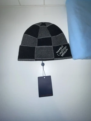 Svartgrå rutig mössa - Snygg svart och grå mössa med broderad text på framsidan. Mössan har ett klassiskt rutmönster och är stickad i mjukt material, perfekt för att hålla värmen och ge en trendig look.