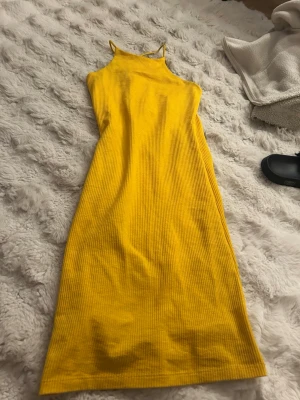 Gul ribbad långklänning - Säljer en snygg gul ribbad långklänning med smala axelband och hög hals. Klänningen har en kroppsnära passform och är perfekt för sommarens alla tillfällen. Materialet är mjukt och stretchigt för en bekväm känsla.
