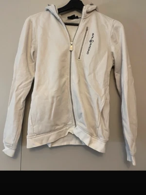 Vit hoodie från Sail Racing - Säljer en vit hoodie från Sail Racing med dragkedja framtill och broderad logga på bröstet. Hoodien har huva och två sidofickor. Tillverkad i mjukt bomullsmaterial som känns skönt mot huden. Perfekt för dig som gillar en clean och sportig stil.