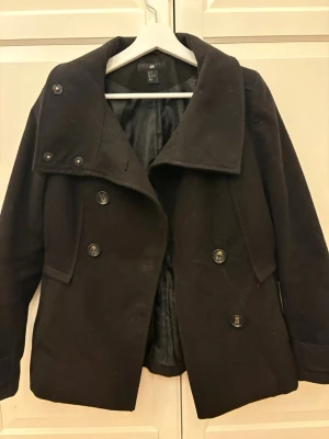 Svart dubbelknäppt kappa - Stilren svart kappa från H&M med dubbelknäppning och stora knappar framtill. Två fickor och kappan har bred krage, dekorativa knappar på axlarna. Fodrad insida och klassisk, rak passform. Perfekt för kyliga dagar.